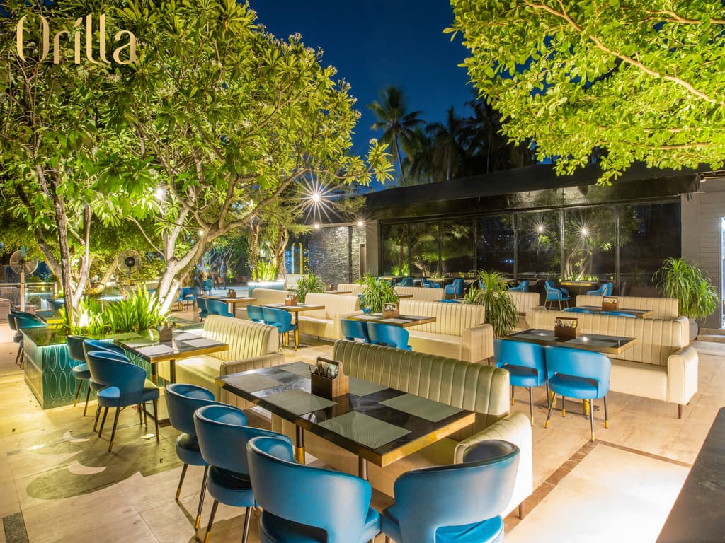 Windy riverside deck with turquoise tub chairs, tan sofas, yellow lighting & lush trees dipping over 